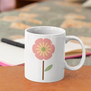 May include: White ceramic mug featuring a pink flower with a yellow centre and a green leaf on a brown stem. The mug is on a wooden table.