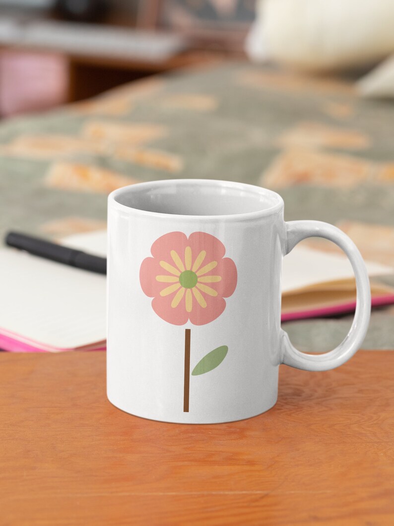 May include: White ceramic mug featuring a pink flower with a yellow centre and a green leaf on a brown stem. The mug is on a wooden table.
