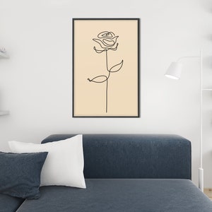 May include: A black framed print of a line drawing of a rose on a beige background. The print is hanging on a white wall above a blue couch with white pillows.