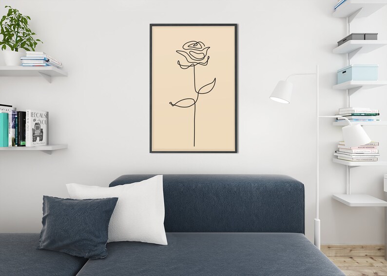 May include: A black framed print of a line drawing of a rose on a beige background. The print is hanging on a white wall above a blue couch with white pillows.