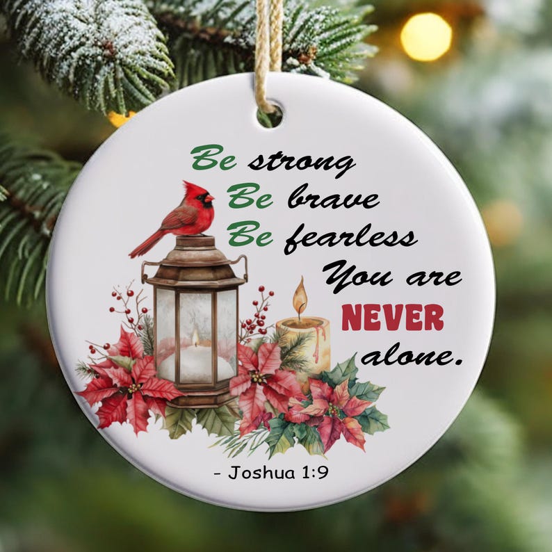 May include: White round Christmas bauble featuring a red cardinal, a lantern, and a candle. The bauble has the text "Be strong, Be brave, Be fearless, You are NEVER alone." and "Joshua 1:9" on it.