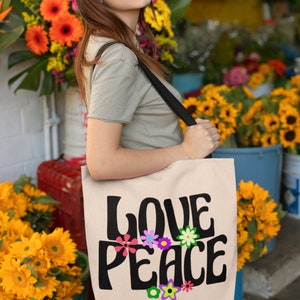 May include: A woman is holding a beige tote bag with a black strap. The bag has the words "LOVE PEACE" printed on it in black with colorful flowers surrounding the text.