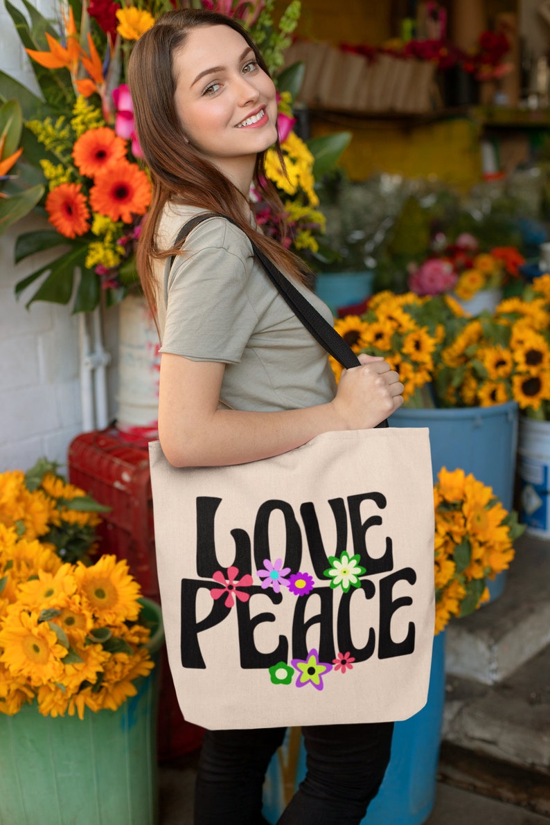 May include: A woman is holding a beige tote bag with a black strap. The bag has the words "LOVE PEACE" printed on it in black with colorful flowers surrounding the text.