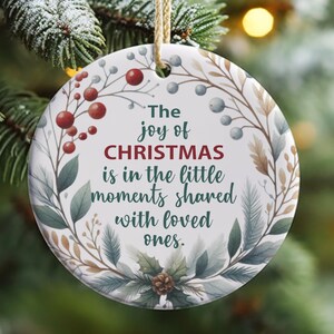 Puede incluir: Un adorno navideño redondo blanco con un diseño floral verde, marrón y rojo. El adorno tiene el texto "The joy of CHRISTMAS is in the little moments shared with loved ones."
