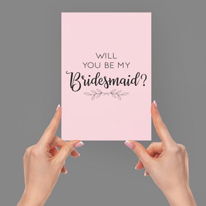 May include: Pink card with the text "Will you be my bridesmaid?" in black cursive font with a small floral design below the text.