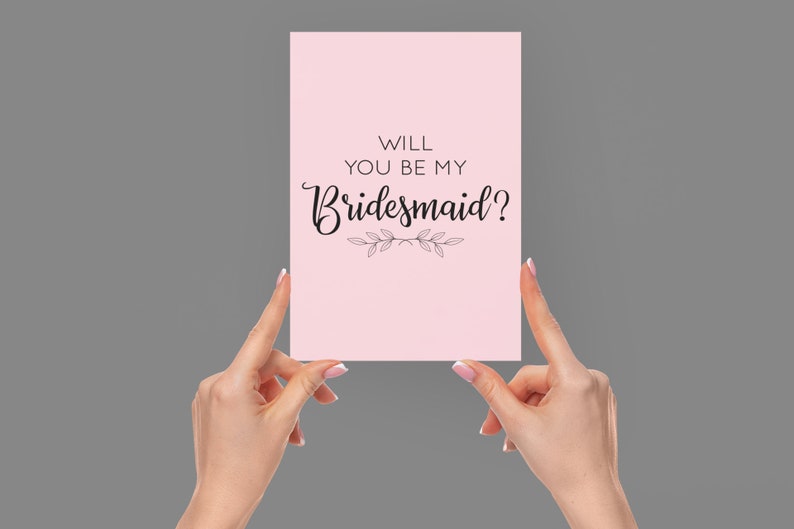 May include: Pink card with the text "Will you be my bridesmaid?" in black cursive font with a small floral design below the text.