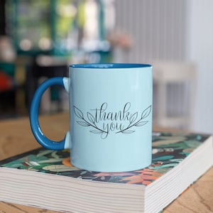 May include: A light blue ceramic mug with a blue handle. The mug has the words "thank you" written in black cursive with a leafy design around the text.