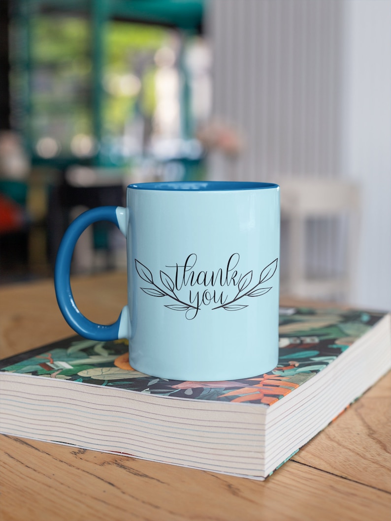 May include: A light blue ceramic mug with a blue handle. The mug has the words "thank you" written in black cursive with a leafy design around the text.