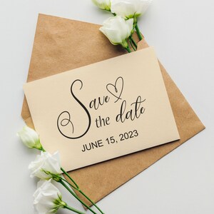 May include: A kraft paper envelope with a cream card inside. The card has a handwritten message that says "Save the date" with a heart and the date "June 15, 2023". White flowers are arranged around the envelope.