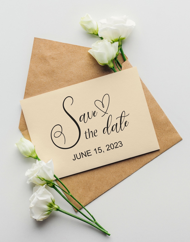 May include: A kraft paper envelope with a cream card inside. The card has a handwritten message that says "Save the date" with a heart and the date "June 15, 2023". White flowers are arranged around the envelope.
