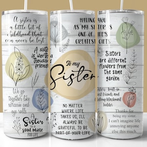 May include: A white tumbler with a straw, featuring a design with the words "To my Sister" and various quotes about sisters. The design includes floral and leaf illustrations in pastel colors, set against a wood-grain background.