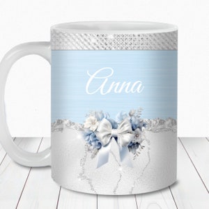 Pode incluir: Uma caneca de cerâmica branca com fundo azul claro e um design de glitter prateado. A caneca tem um laço branco com um design de glitter prateado e o nome "Anna" escrito em itálico branco.