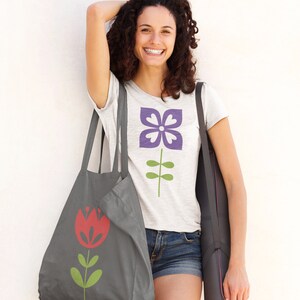 May include: A woman is carrying a grey tote bag with a red flower design and a black yoga mat. She is wearing a white t-shirt with a purple flower design and denim shorts.