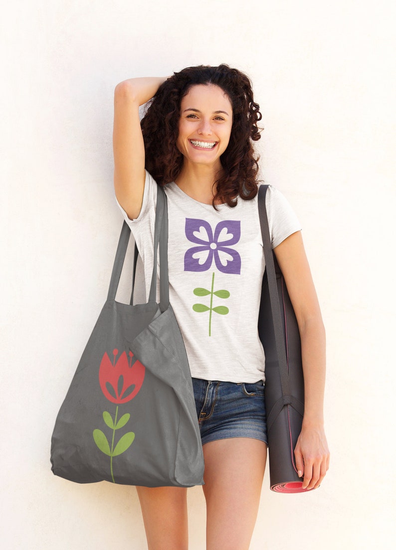 May include: A woman is carrying a grey tote bag with a red flower design and a black yoga mat. She is wearing a white t-shirt with a purple flower design and denim shorts.