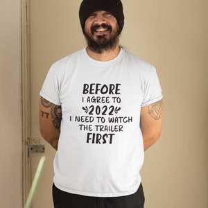 May include: A white t-shirt with black text that reads "Before I agree to 2022 I need to watch the trailer first". A man is wearing the shirt and smiling at the camera.