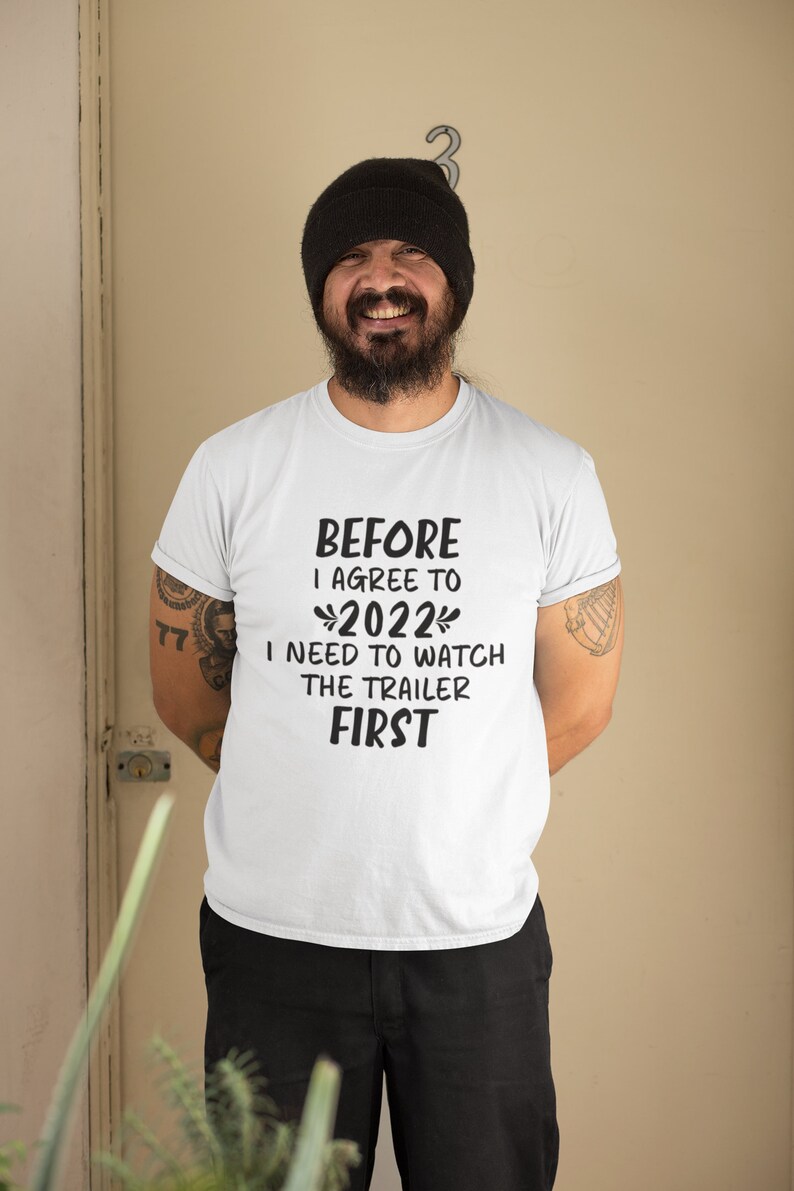 May include: A white t-shirt with black text that reads "Before I agree to 2022 I need to watch the trailer first". A man is wearing the shirt and smiling at the camera.