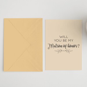May include: A beige card with black text that reads "Will you be my Matron of honor?" with a small floral design below the text. The card is sitting on a white surface next to a beige envelope.