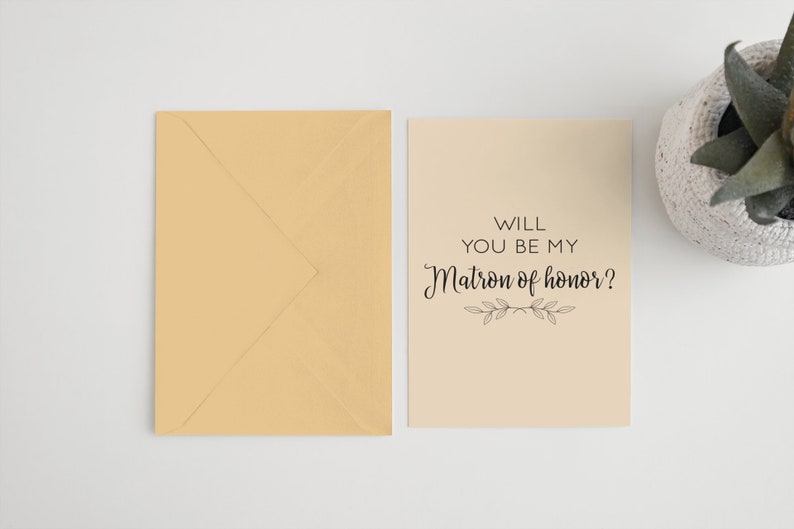 May include: A beige card with black text that reads "Will you be my Matron of honor?" with a small floral design below the text. The card is sitting on a white surface next to a beige envelope.