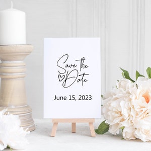 May include: White card with black calligraphy that says "Save the Date" with a small heart symbol and the date June 15, 2023. The card is on a small wooden easel.