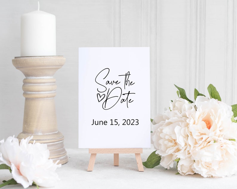 May include: White card with black calligraphy that says "Save the Date" with a small heart symbol and the date June 15, 2023. The card is on a small wooden easel.