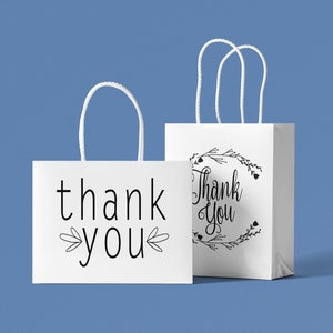 May include: Two white paper gift bags with black handles. One bag says "thank you" with a simple floral design. The other bag says "Thank You" with a more elaborate floral design.