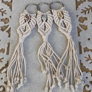 Puede incluir: Tres llaveros de macramé blanco con abalorios de concha dorada y plateada. Cada llavero tiene un anillo plateado y una borla larga.