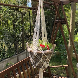 Peut inclure: Un porte-plante en macramé blanc avec un panier en osier tissé rempli de fleurs roses et rouges. Le porte-plante est suspendu à une structure en bois.