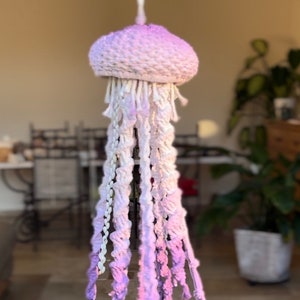 May include: A crocheted jellyfish mobile with a purple and white ombre color scheme. The jellyfish is hanging from a white cord.