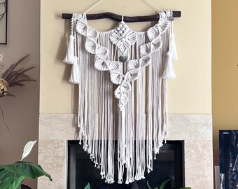 May include: A cream-colored macrame wall hanging suspended from a dark brown wooden branch. The design features intricate knotted leaf-like patterns, a central stone accent, and long fringe ending in tassels on both sides with a white hanging cord.