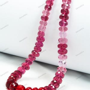 May include: A necklace composed of faceted, reddish-pink gemstones. The beads transition in color from deep red to lighter pink hues. The necklace is displayed against a white background, showcasing the varying shades and the polished surfaces of the stones.