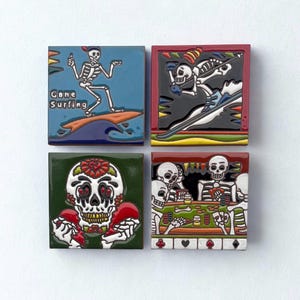 May include: Four square ceramic tiles with Day of the Dead skeleton designs. One shows a skeleton surfing with the text "Gone Surfing". Other tiles depict skeletons skiing, with a heart, and playing poker. Each tile has a different color border.