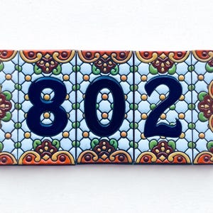 May include: Rectangular ceramic tile displaying the house number 802 in navy blue. The tile has a light blue background with a repeating pattern of orange, green, and yellow floral designs. The tile is rectangular.