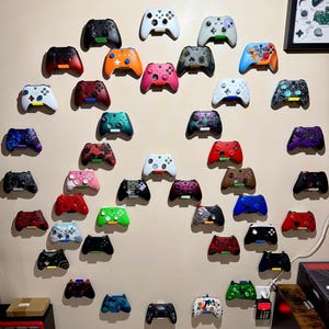 May include: A collection of colorful video game controllers mounted on a wall. The controllers feature a variety of colors, including black, white, red, blue, green, and pink. Some controllers have unique designs and patterns.