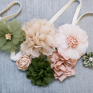 May include: A white satin ribbon with a variety of fabric flowers in shades of green, beige, and pink. The flowers are arranged in a cluster and are attached to the ribbon.