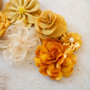 K&ouml;nnte beinhalten: Eine Sammlung von gelben und goldenen Stoffblumen. Die Blumen sind aus verschiedenen Materialien gefertigt, darunter Seide, Satin und T&uuml;ll. Sie sind in einer Gruppe auf einem wei&szlig;en Hintergrund angeordnet.