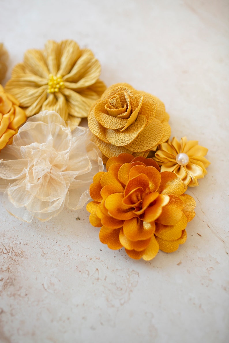 K&ouml;nnte beinhalten: Eine Sammlung von gelben und goldenen Stoffblumen. Die Blumen sind aus verschiedenen Materialien gefertigt, darunter Seide, Satin und T&uuml;ll. Sie sind in einer Gruppe auf einem wei&szlig;en Hintergrund angeordnet.