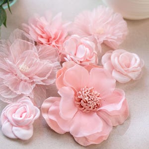 Peut inclure: Un assortiment de fleurs en tissu rose, comprenant des roses et de plus grandes fleurs aux pétales détaillés. Les fleurs sont disposées sur une surface claire, mettant en valeur leurs textures délicates et nuances de rose.