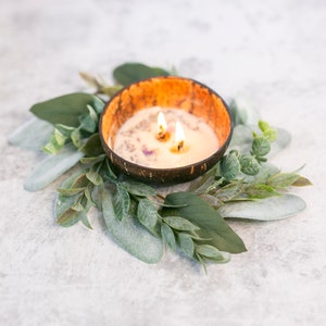 Puede incluir: Una vela blanca con tres mechas ardiendo en un cuenco de c&aacute;scara de coco natural. El cuenco est&aacute; rodeado por una corona de hojas verdes.