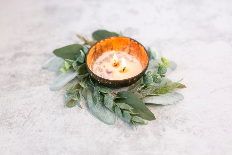 Puede incluir: Una vela blanca con tres mechas ardiendo en un cuenco de c&aacute;scara de coco natural. El cuenco est&aacute; rodeado por una corona de hojas verdes.