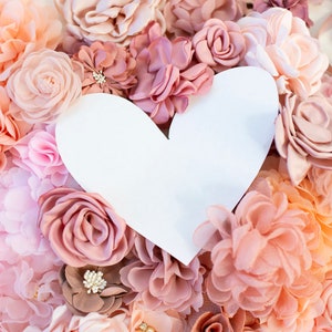 Puede incluir: Un coraz&oacute;n de papel blanco recortado rodeado de una variedad de flores de tela rosa y melocot&oacute;n. Las flores est&aacute;n dispuestas en un patr&oacute;n circular, creando un fondo rom&aacute;ntico y delicado para el coraz&oacute;n.