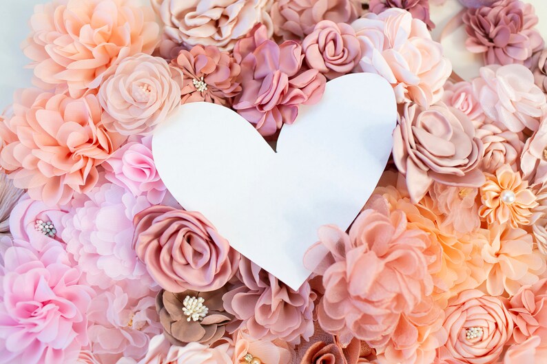 Puede incluir: Un coraz&oacute;n de papel blanco recortado rodeado de una variedad de flores de tela rosa y melocot&oacute;n. Las flores est&aacute;n dispuestas en un patr&oacute;n circular, creando un fondo rom&aacute;ntico y delicado para el coraz&oacute;n.