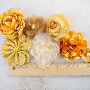 K&ouml;nnte beinhalten: Sortiment aus sechs gelben Stoffblumen, darunter eine Rose, eine G&auml;nsebl&uuml;mchen und eine Chrysantheme. Die Blumen sind auf einer wei&szlig;en Oberfl&auml;che mit einem Holzlineal zum Ma&szlig;stab angeordnet.