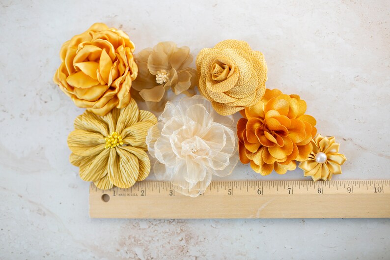 K&ouml;nnte beinhalten: Sortiment aus sechs gelben Stoffblumen, darunter eine Rose, eine G&auml;nsebl&uuml;mchen und eine Chrysantheme. Die Blumen sind auf einer wei&szlig;en Oberfl&auml;che mit einem Holzlineal zum Ma&szlig;stab angeordnet.