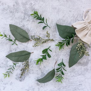 May include: A white hessian bag with a drawstring closure, surrounded by green leaves and sprigs on a grey background.