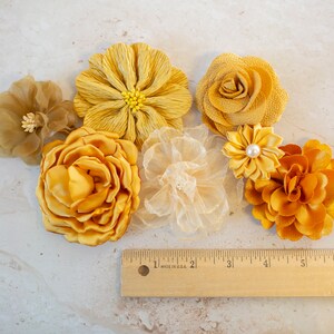 K&ouml;nnte beinhalten: Sortiment aus sechs gelben Stoffblumen, darunter eine gro&szlig;e, gekr&auml;uselte Blume, eine kleinere, zierlichere Blume und ein paar kleinere, kompaktere Blumen. Die Blumen sind auf einer hellbeigen Oberfl&auml;che mit einem Holzlineal angeordnet.