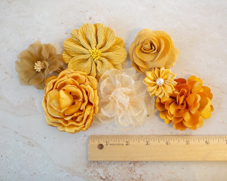 K&ouml;nnte beinhalten: Sortiment aus sechs gelben Stoffblumen, darunter eine gro&szlig;e, gekr&auml;uselte Blume, eine kleinere, zierlichere Blume und ein paar kleinere, kompaktere Blumen. Die Blumen sind auf einer hellbeigen Oberfl&auml;che mit einem Holzlineal angeordnet.