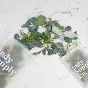 May include: Two clear plastic bags with the text "Indy Supply Co." printed on them. The bags are filled with a variety of green leaves and sprigs.