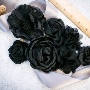 May include: A collection of six black fabric flowers, including a large anemone, a rose, a smaller rose, a ranunculus, and two smaller flowers. The flowers are arranged on a silver ribbon.