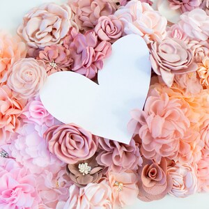 Puede incluir: Un recorte de papel blanco en forma de coraz&oacute;n rodeado de una variedad de flores de tela rosa, melocot&oacute;n y marr&oacute;n.