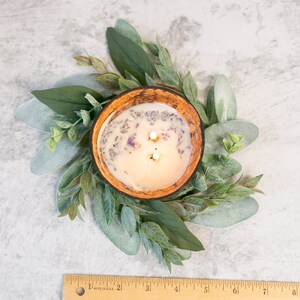 Puede incluir: Una vela de dos mechas en un cuenco de madera con flores de lavanda secas y una corona de eucalipto verde alrededor.
